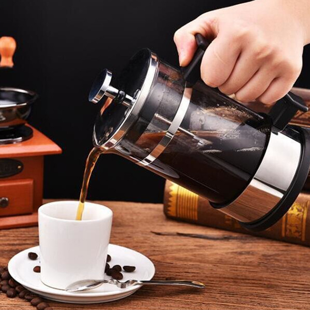 Eine Hand gießt Kaffee aus einer French Press in eine weiße Tasse auf einer Untertasse. Kaffeebohnen und ein Löffel sind in der Nähe der Tasse.