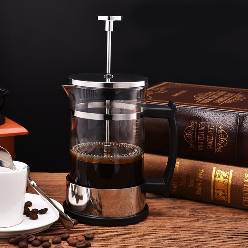 Eine French Press mit Kaffee steht auf einem Holztisch, neben Kaffeebohnen, einer Tasse und Büchern.