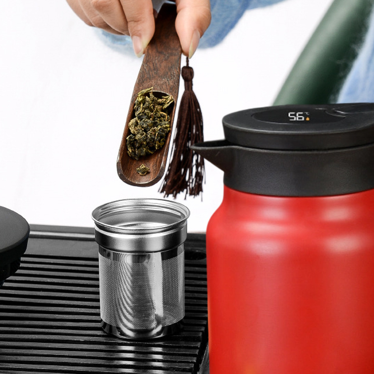 Eine Hand schüttet Teeblätter von einem Holzlöffel in einen Metall-Tee-Infuser, in der Nähe eines roten Wasserkochers.