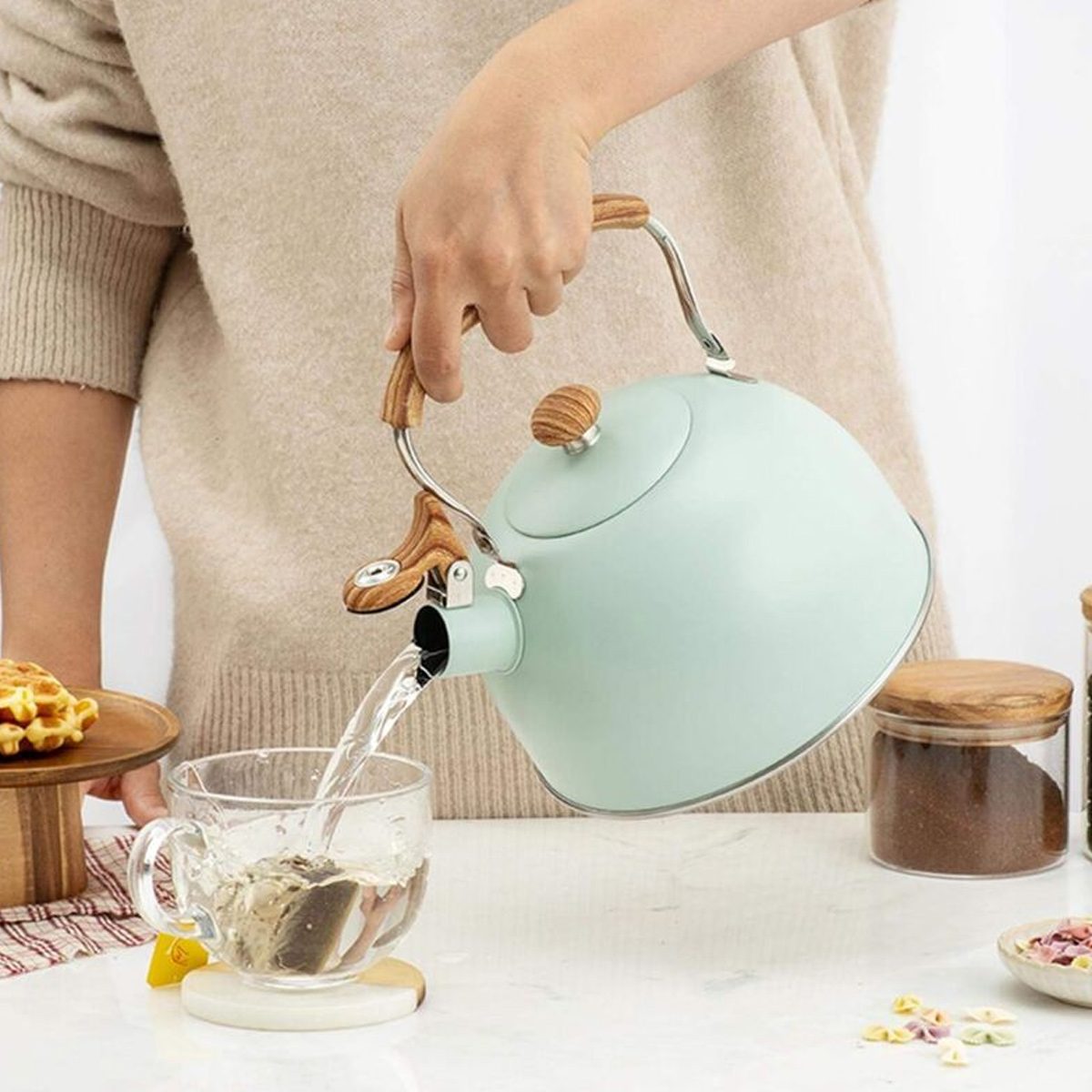 Eine Person gießt heißes Wasser aus einem blauen Wasserkocher in eine Glastasse mit Teebeutel auf einem weißen Tisch. Holzakzente sind sichtbar.