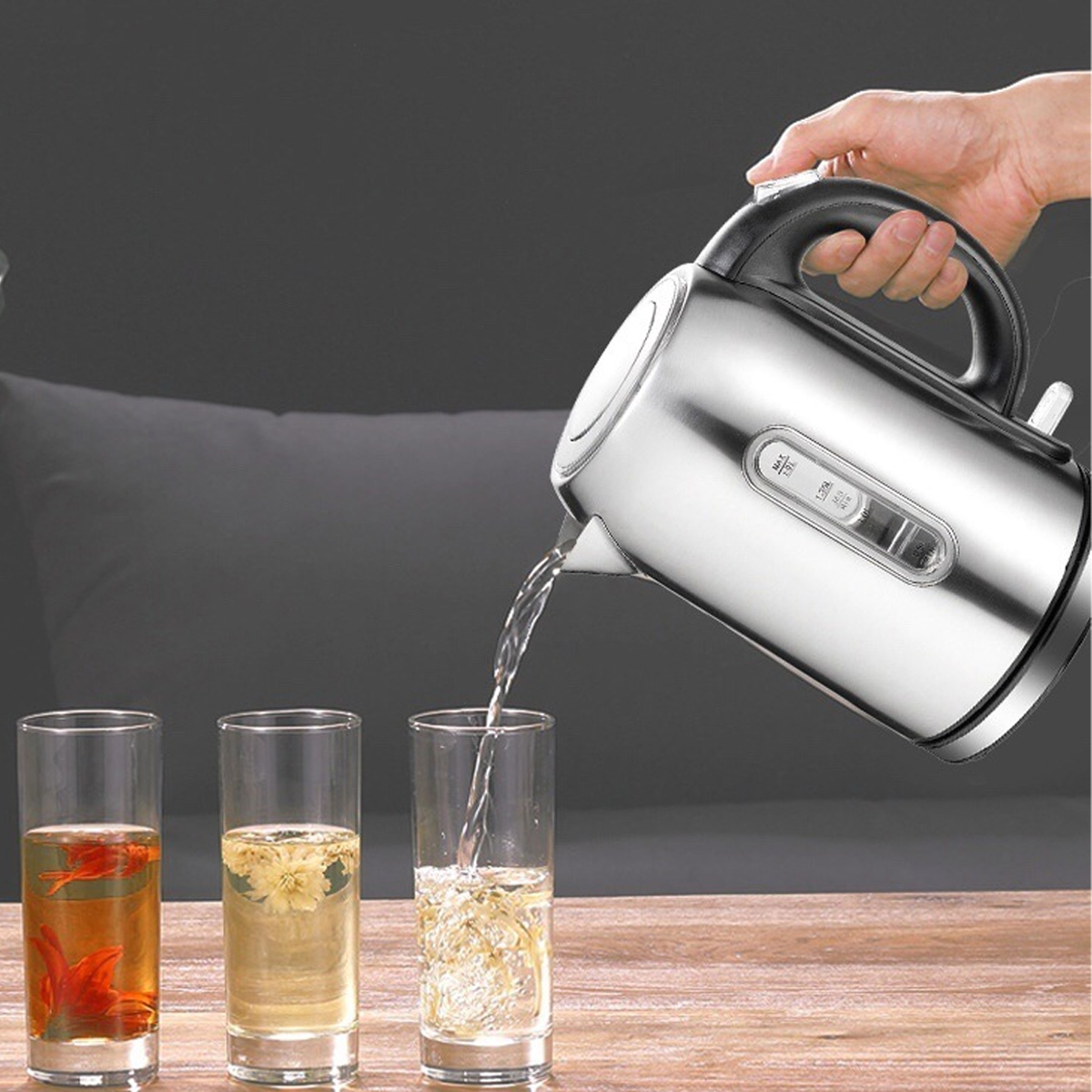 Eine Hand gießt Wasser aus einem silbernen Wasserkocher in ein Glas. Drei Gläser stehen auf einem Tisch, jedes mit unterschiedlichem Tee und Blumen.