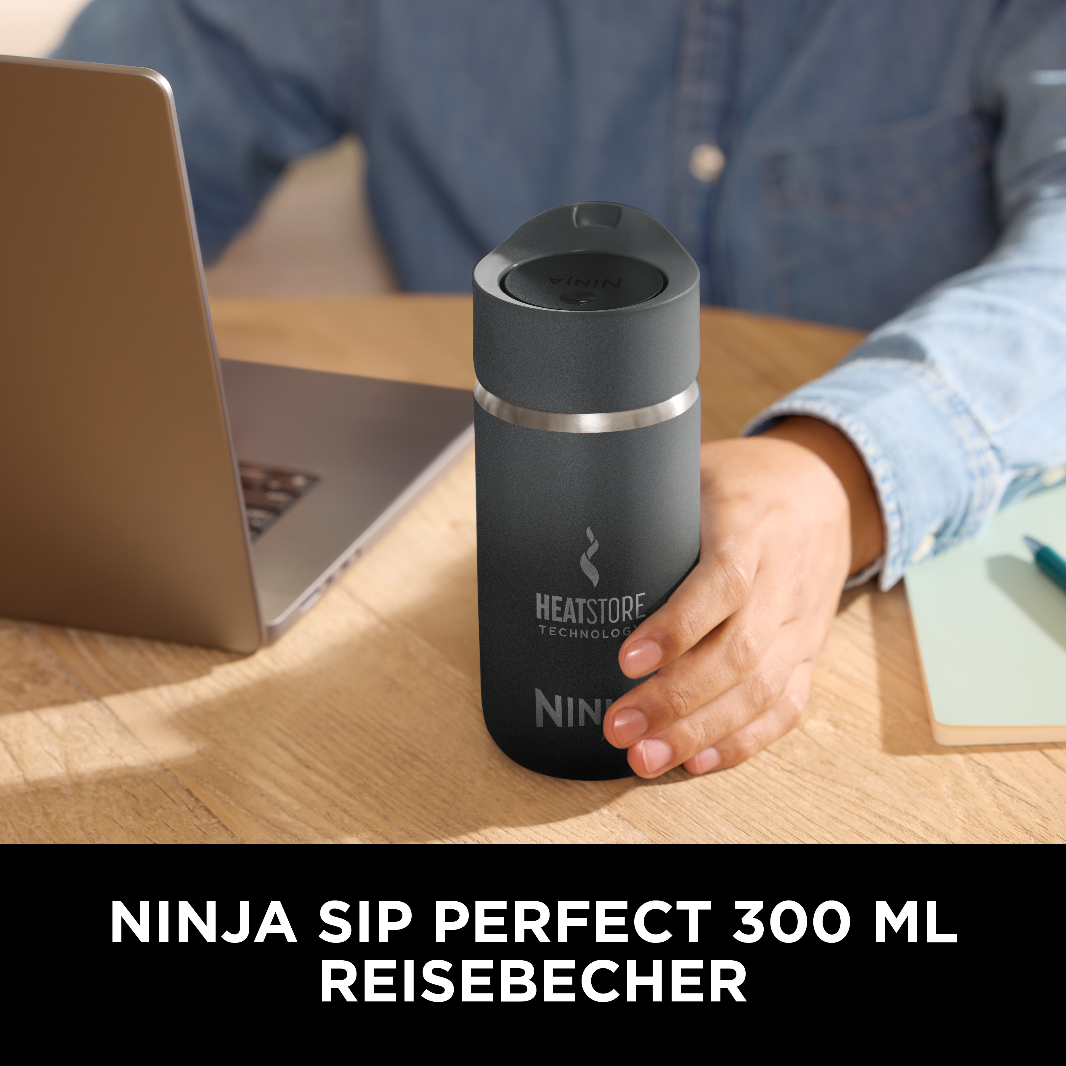 Una persona tiene in mano un bicchiere da viaggio Ninja nero. Un laptop e un quaderno sono su una scrivania di legno.