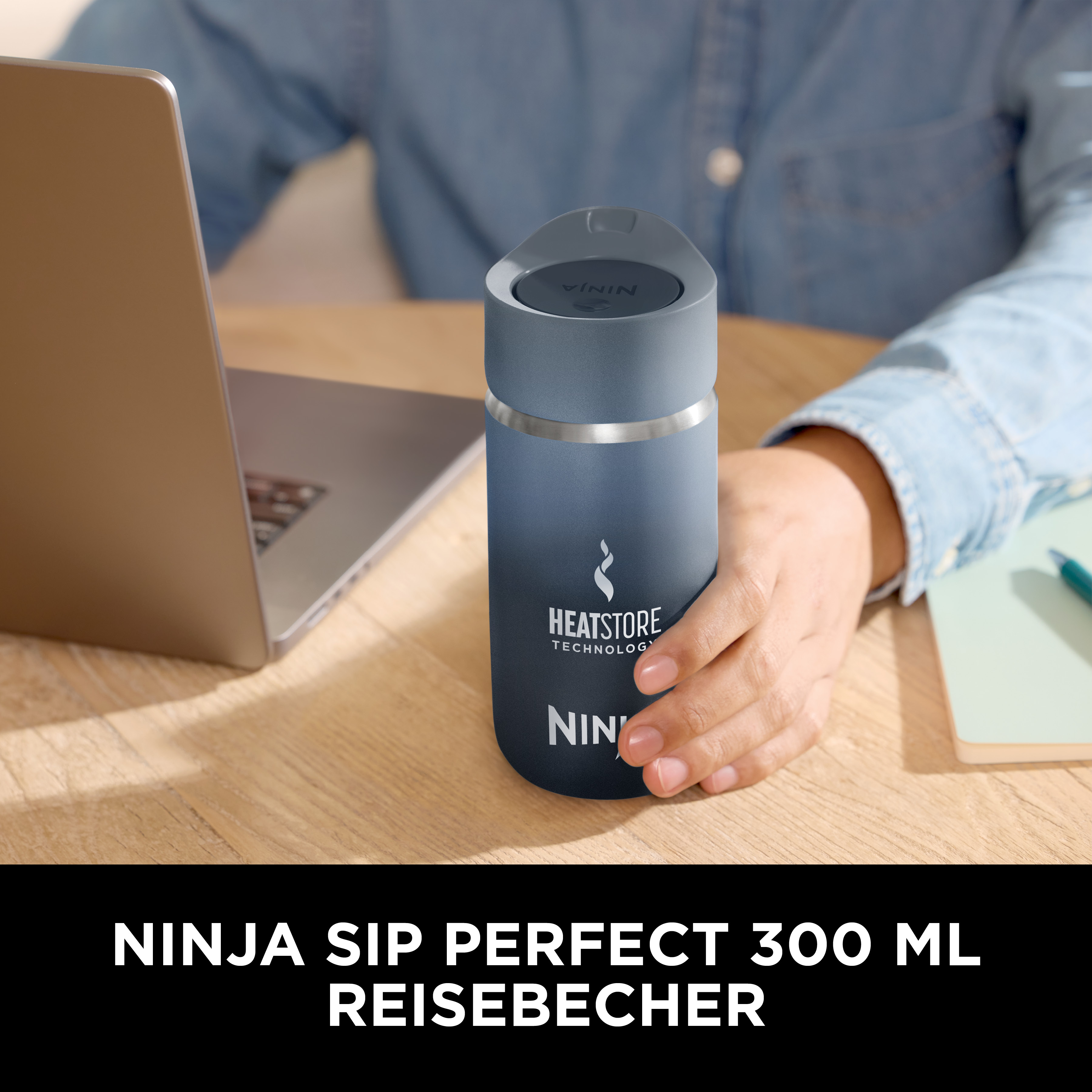 Una persona tiene in mano un bicchiere da viaggio Ninja blu. Un laptop e un taccuino sono su una scrivania di legno. Testo: Ninja Sip Perfect.