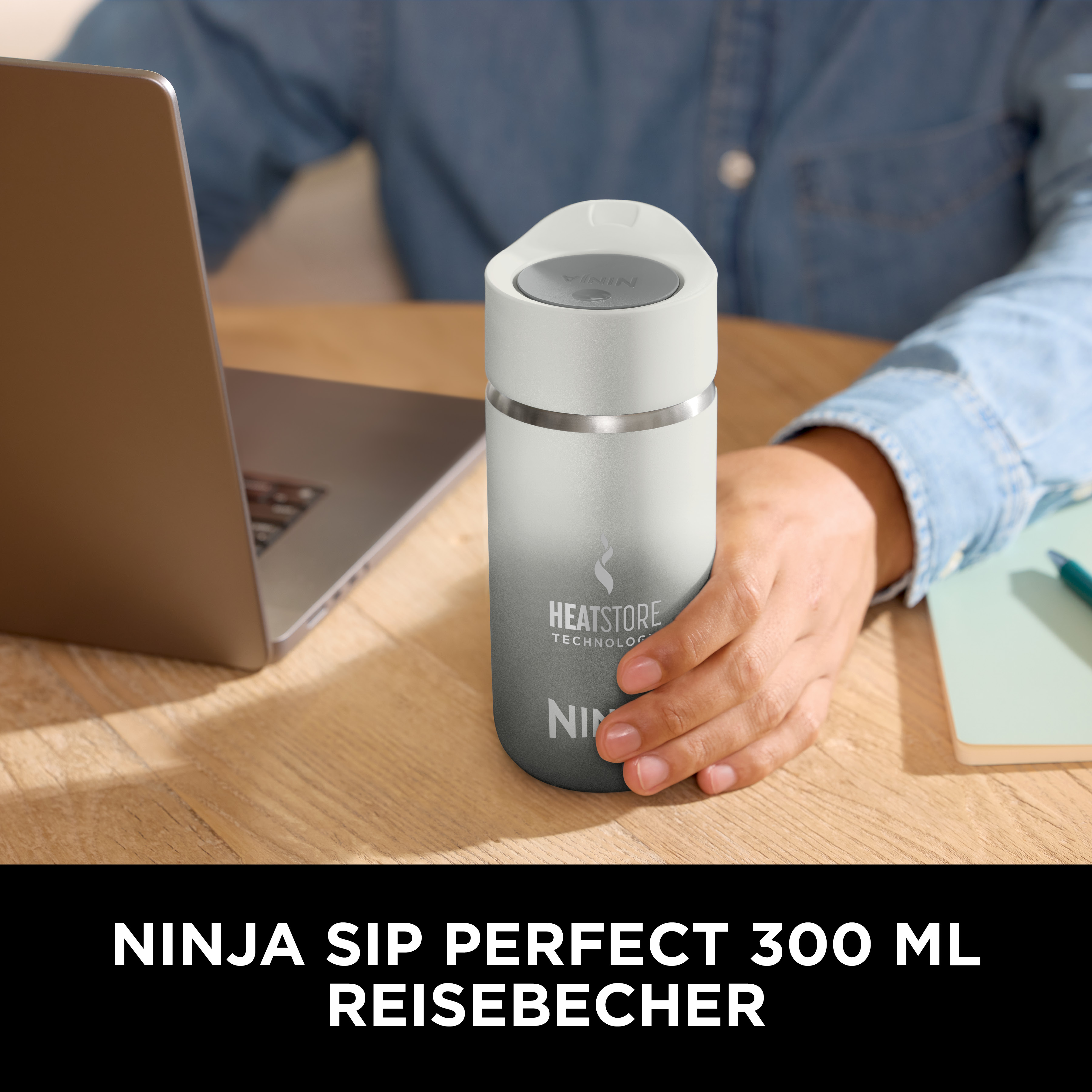 Une personne tient un mug de voyage blanc et gris près d'un ordinateur portable. Le mug a une marque. Un carnet et un stylo sont également présents.