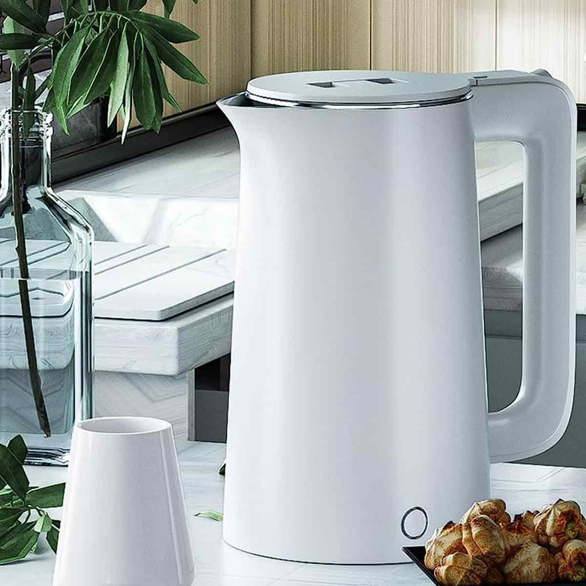 Ein weißer Wasserkocher steht auf einer Theke. Eine Vase mit einer Pflanze, eine weiße Tasse und ein Teller mit Essen daneben.