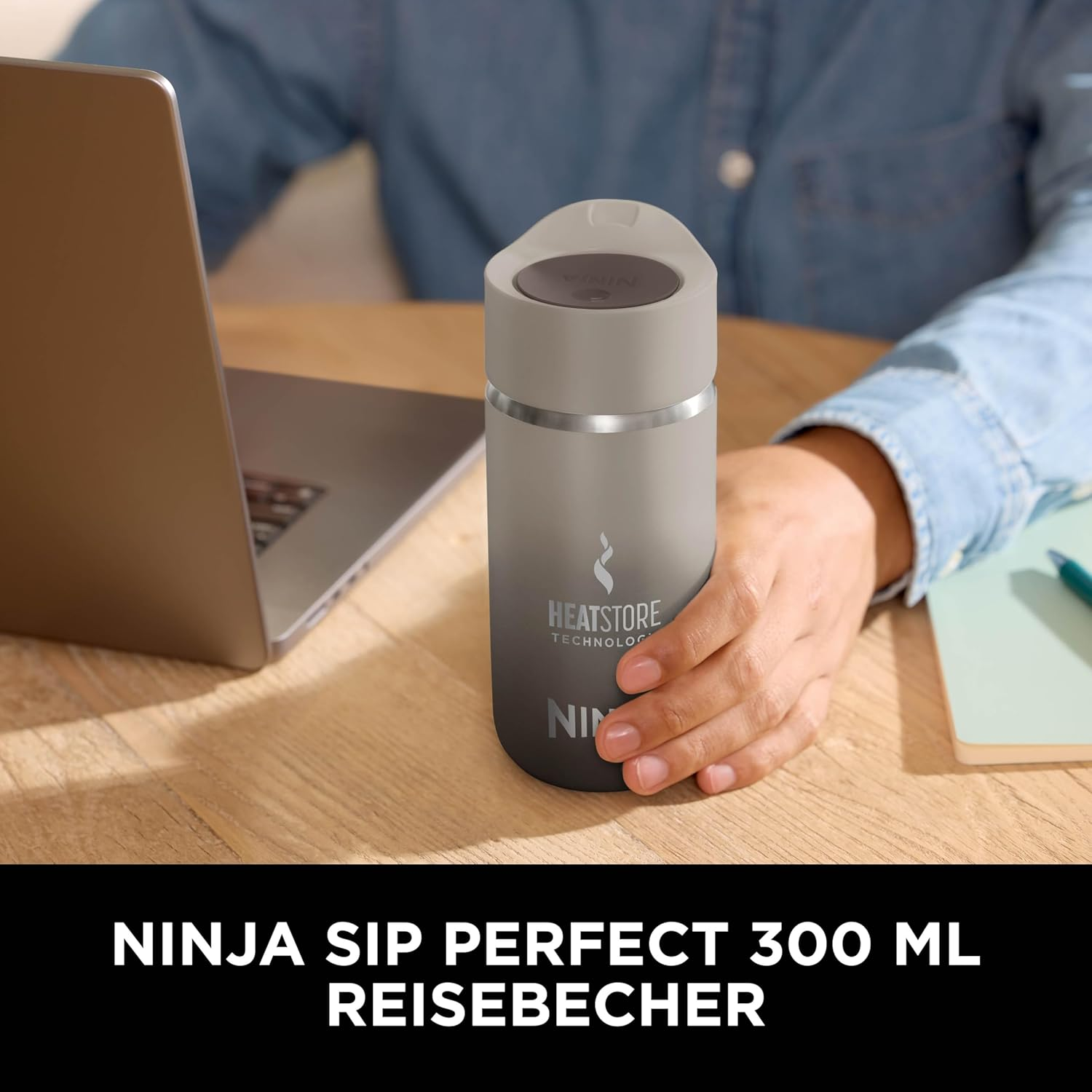 Une personne tient un mug de voyage Ninja sur une table en bois. Un ordinateur portable et un carnet sont également sur la table. Le mug indique 'Heatstore Technology'.