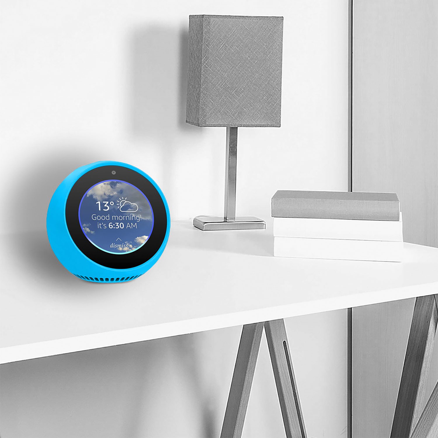 Ein blaues Smart-Home-Gerät auf einem weißen Tisch. Eine graue Lampe und Bücher dahinter. Der Bildschirm zeigt Wetter und Uhrzeit.