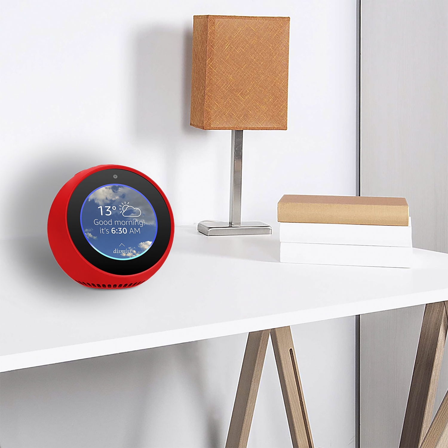Un dispositivo smart home rosso mostra il meteo. Una lampada e libri sono su un tavolo bianco. Parete bianca sullo sfondo.