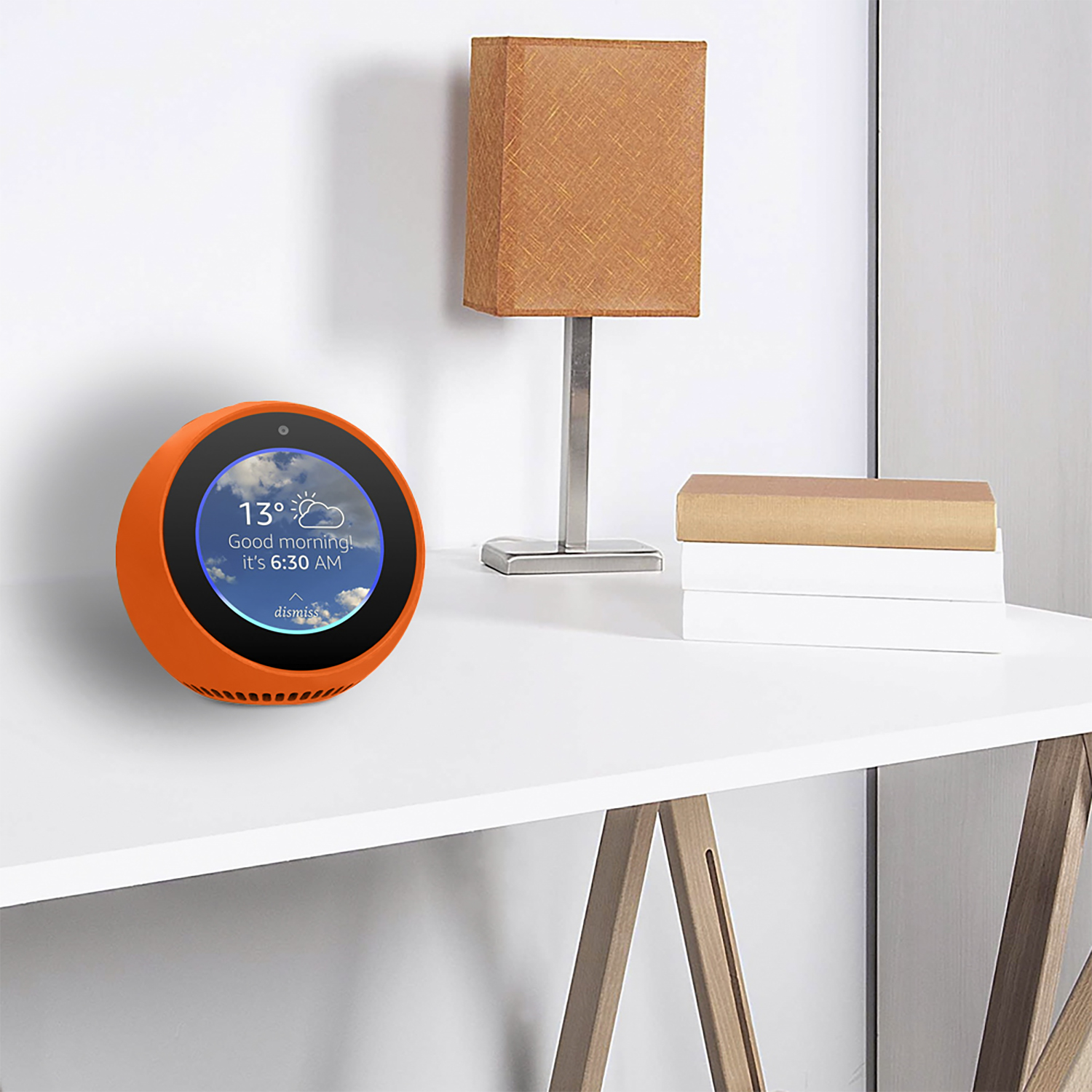 Un altoparlante intelligente arancione mostra meteo e ora. Una lampada e libri sono su un tavolo bianco. Parete bianca sullo sfondo.