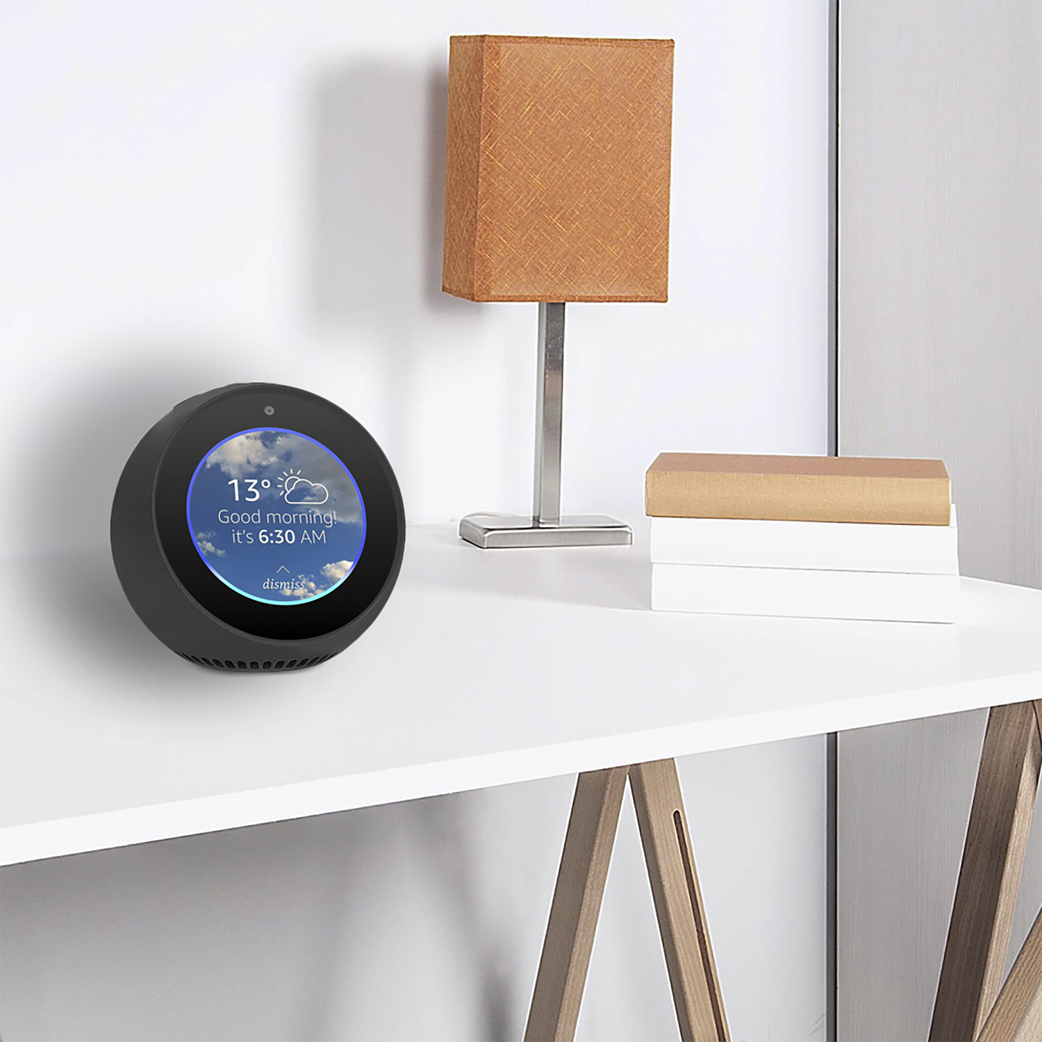 Un altoparlante intelligente con schermo che mostra informazioni meteo è su un tavolo bianco. Una lampada e dei libri sullo sfondo.