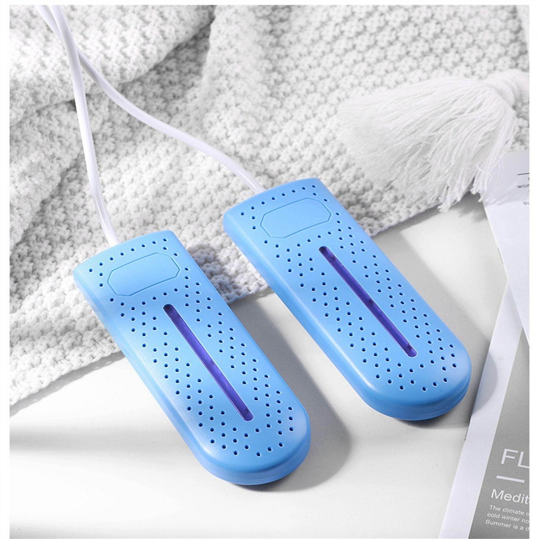 Zwei blaue Schuhtrockner mit weißen Kabeln auf einer weißen Strickdecke und einer weißen Quaste.