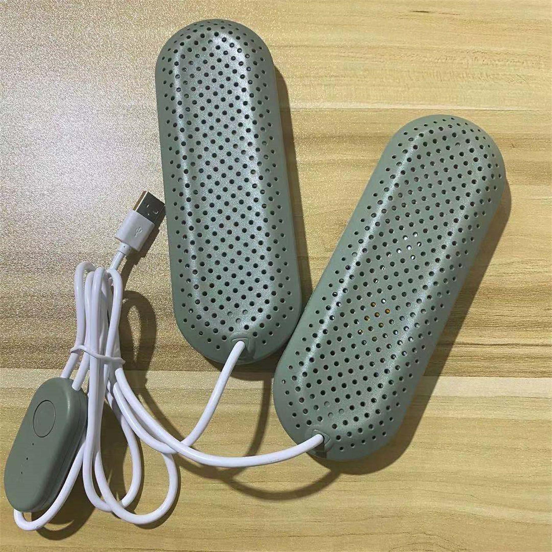 Zwei graue Schuhtrockner mit perforierten Oberflächen, verbunden mit einem weißen USB-Kabel und einem Steuerschalter, auf einer Holzoberfläche.