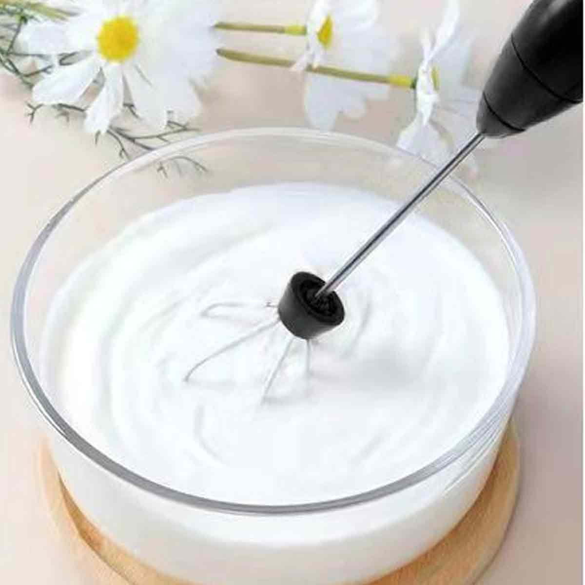 Ein Handmixer in einer Schüssel mit weißer Sahne. Weiße Blumen sind im Hintergrund. Die Schüssel steht auf einem Holzsockel.