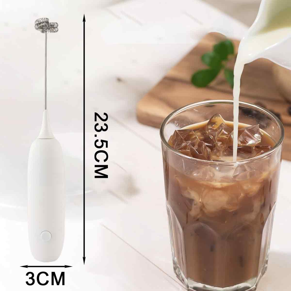 Ein weißer Milchaufschäumer, ein Glas Eiskaffee wird mit Milch übergossen. Ein Holzbrett mit einer Pflanze im Hintergrund.