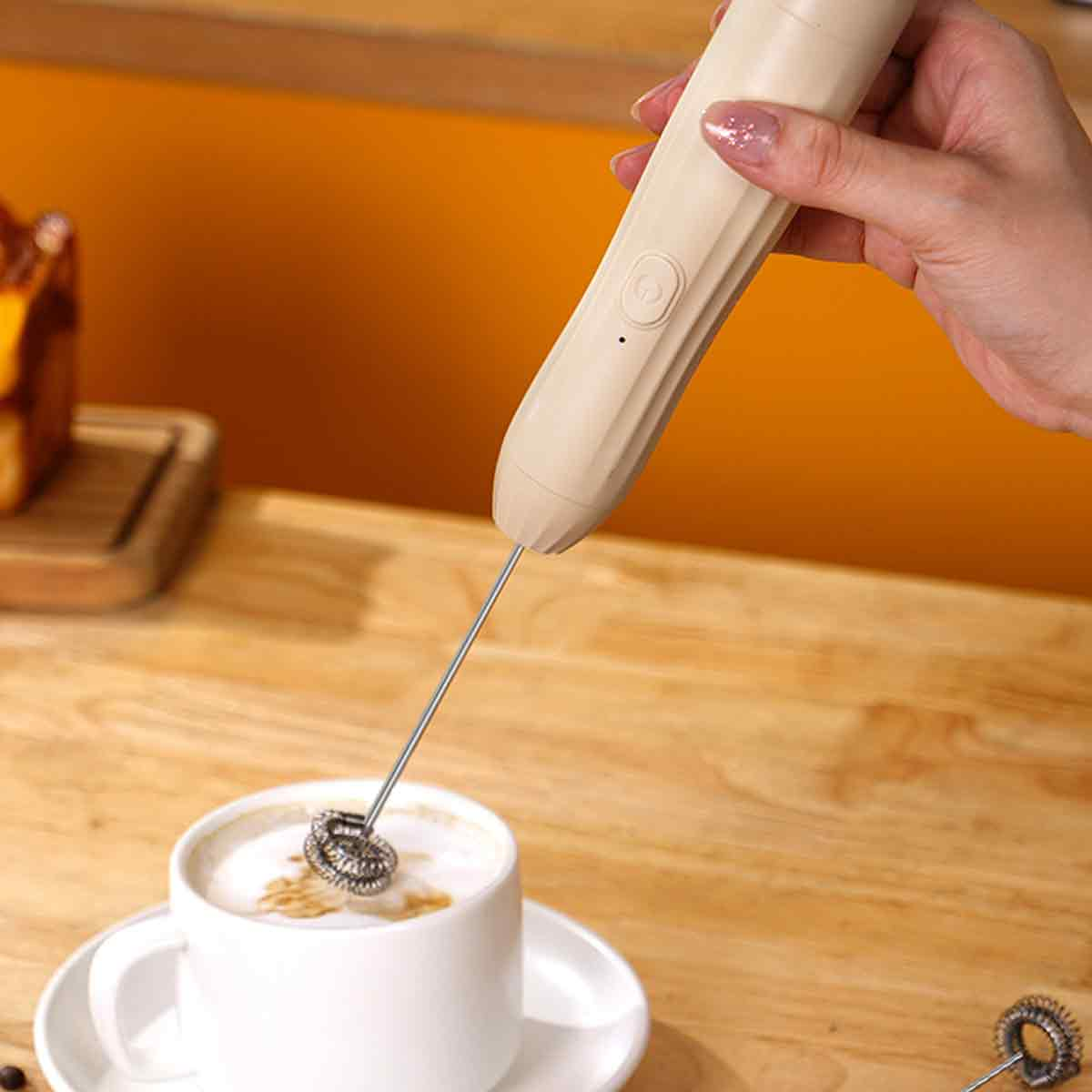 Eine Hand hält einen beigefarbenen Milchaufschäumer über einer weißen Kaffeetasse mit Untertasse. Der Hintergrund ist eine orangefarbene Wand.