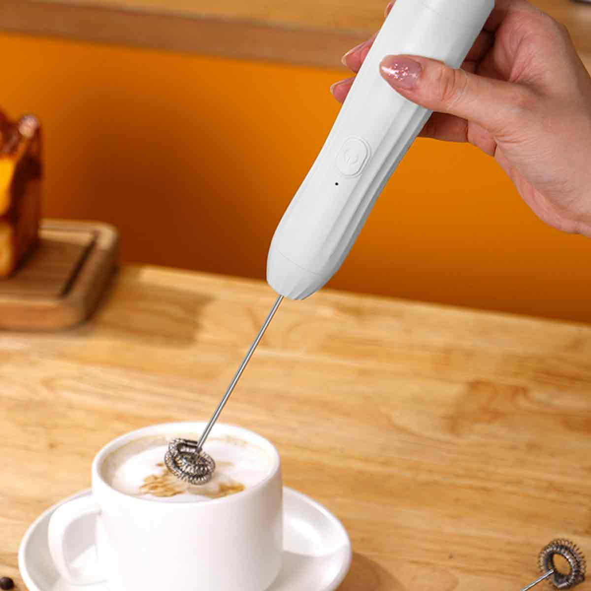 Eine Hand hält einen weißen Milchaufschäumer über einer Tasse Kaffee mit Schaum. Im Hintergrund ist ein Kuchen.