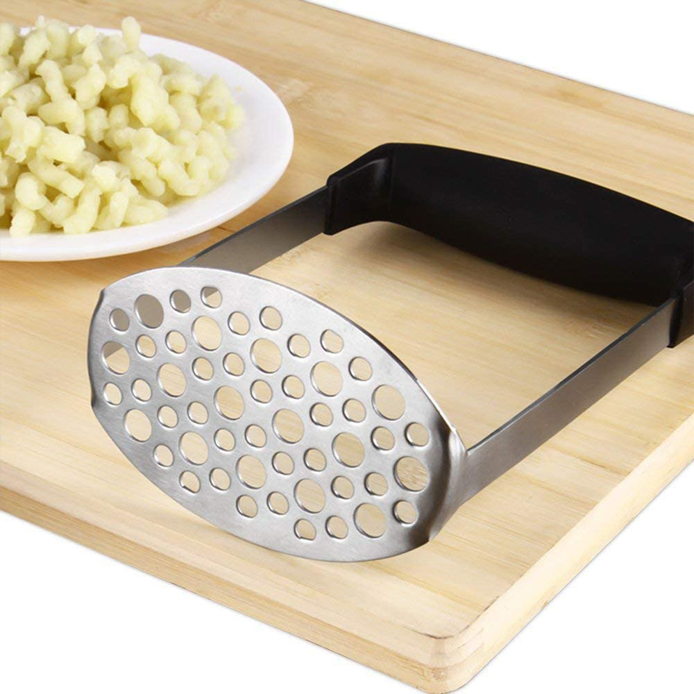 Ein Kartoffelstampfer aus Metall mit schwarzem Griff liegt auf einem Holzbrett neben einem Teller mit Knödeln.