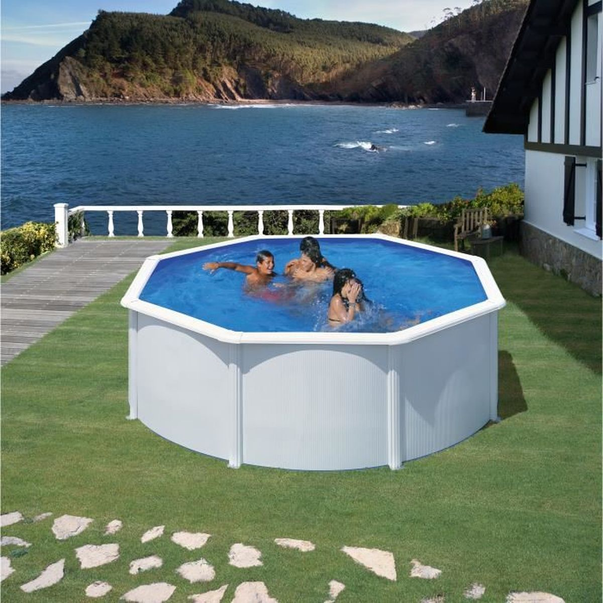 Tres personas nadan en una piscina azul. La piscina está en un jardín verde, cerca del mar y una casa.