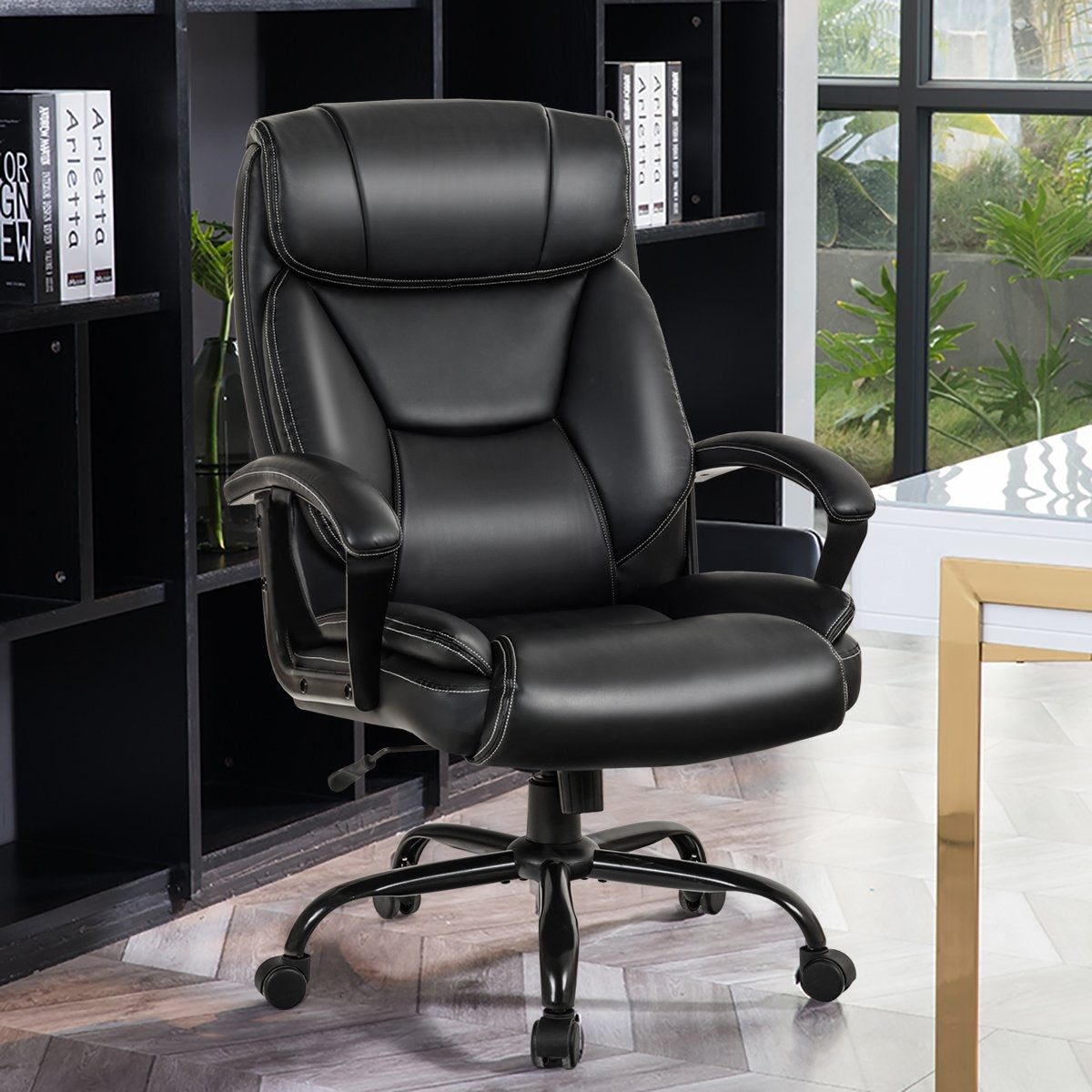 Una silla de oficina de cuero negro con costuras blancas. Tiene respaldo alto, reposabrazos y base de cinco ruedas. Estanterías y una ventana al fondo.
