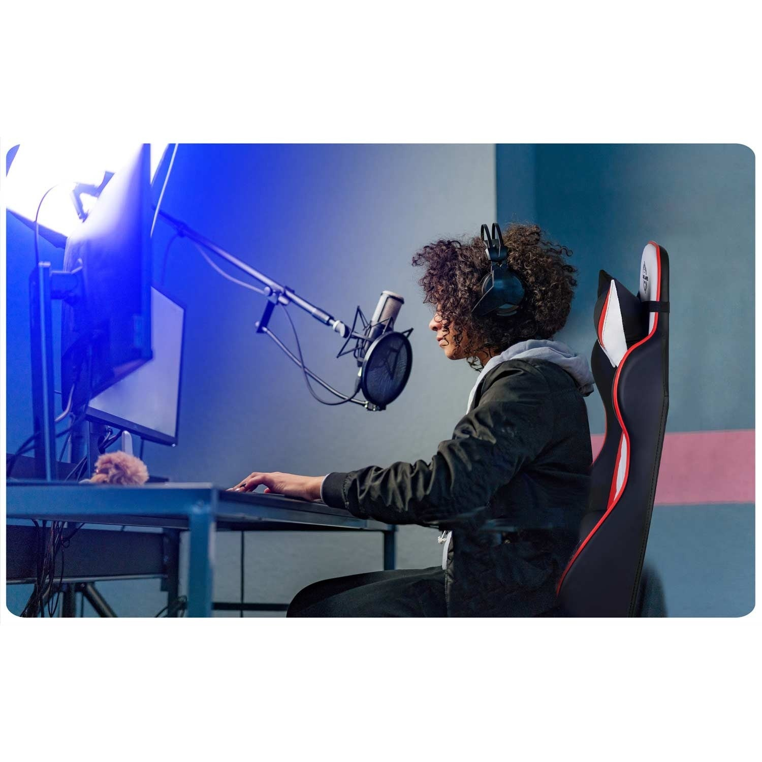 Una persona con auriculares está sentada en una silla de juego, usando una computadora. Hay un micrófono delante. El fondo es un estudio.