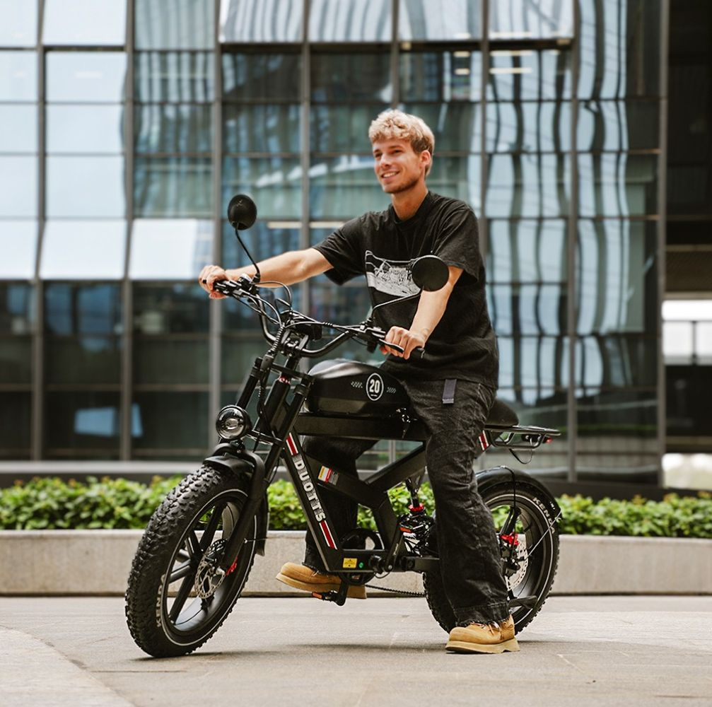 Un uomo su una bici elettrica nera sorride. Indossa una camicia nera e jeans. Sullo sfondo ci sono edifici e cespugli verdi.