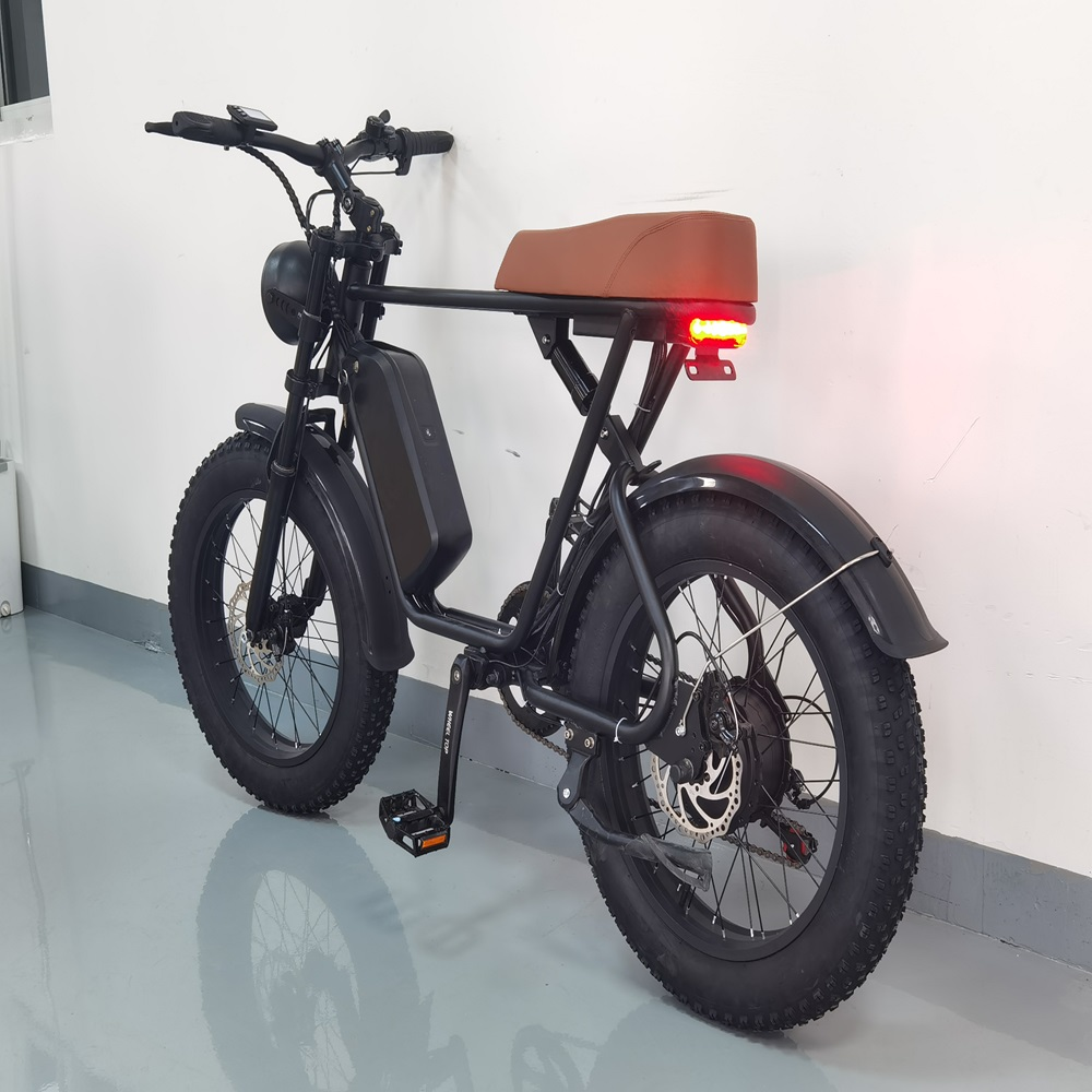 Bicicletta elettrica nera con sella marrone e luce posteriore rossa, parcheggiata contro un muro bianco su un pavimento riflettente.