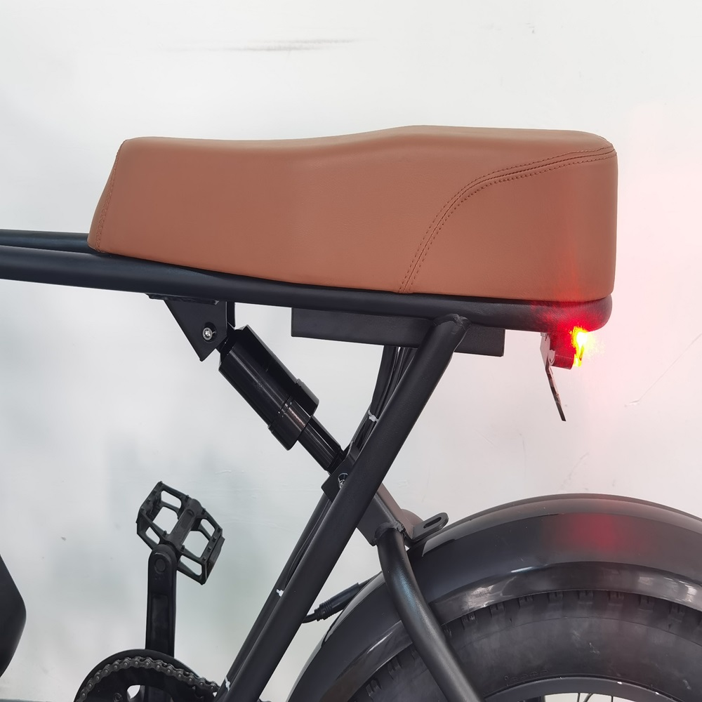 Una sella da bici in pelle marrone è su un telaio nero. Sono visibili il pneumatico posteriore e la luce posteriore rossa. È presente un pedale nero.