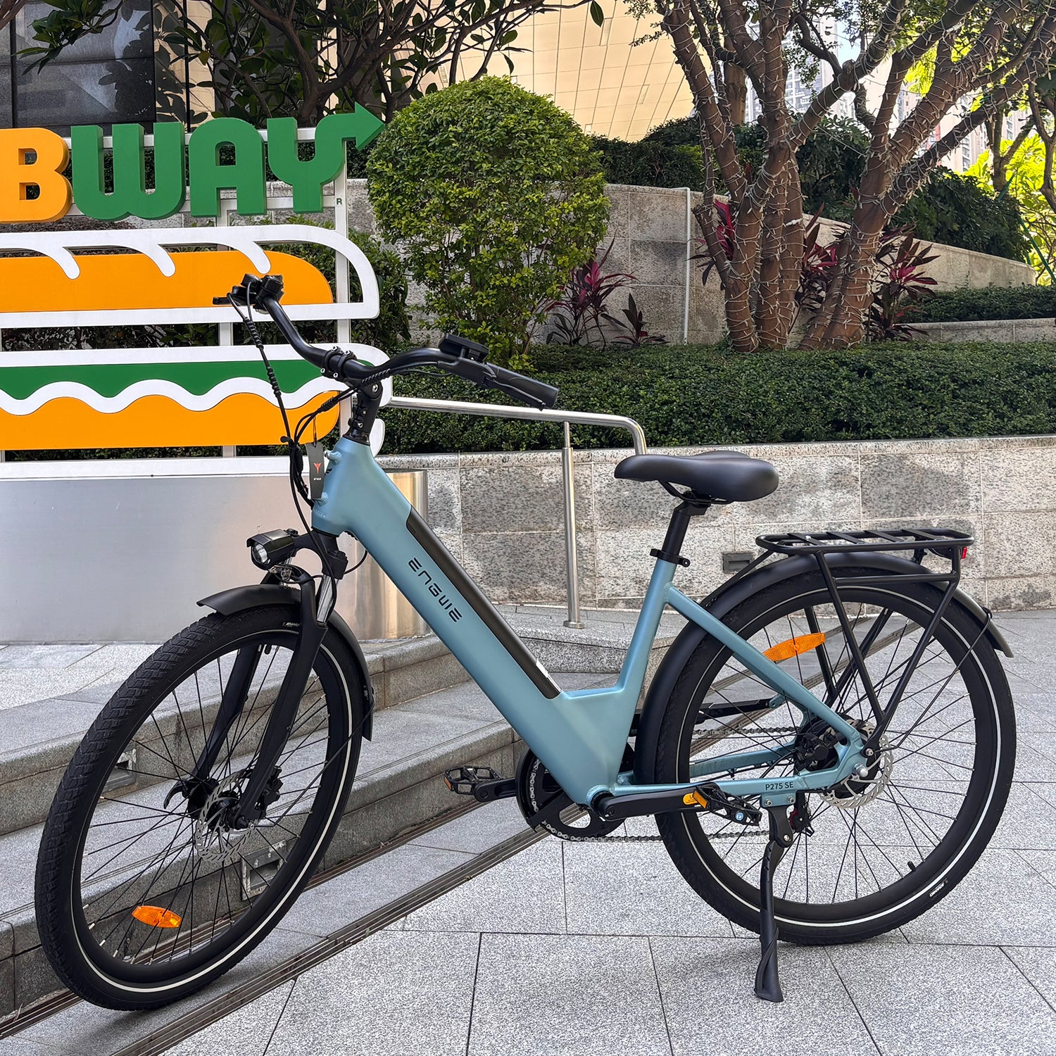 Una bicicleta eléctrica azul claro estacionada frente a un letrero de Subway. La bicicleta está sobre una superficie embaldosada cerca de un seto verde.
