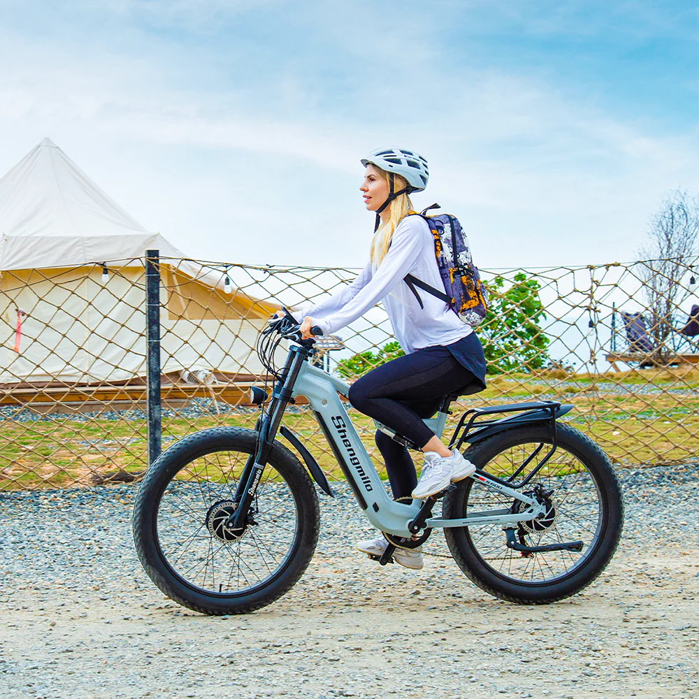 Donna su e-bike grigia con casco bianco. Tenda e recinzione sullo sfondo. Strada sterrata. Cielo blu nuvoloso.