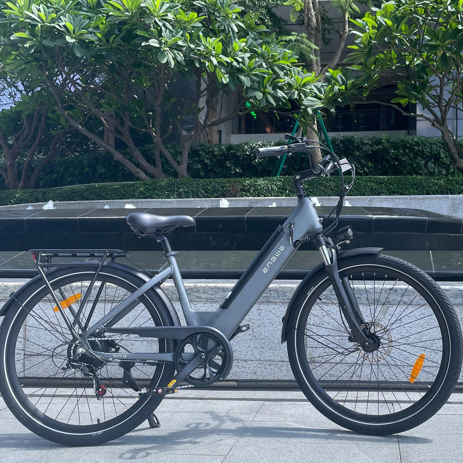 Una e-bike ENGWE gris está estacionada en una acera. Árboles y una fuente en el fondo.