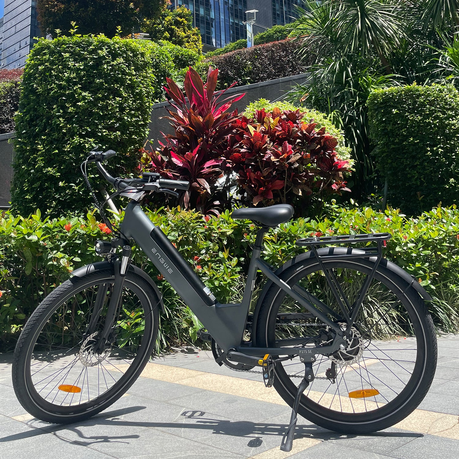 Una bicicleta eléctrica está estacionada en una acera. Hay vegetación exuberante y un edificio en el fondo. La bicicleta es gris con neumáticos negros.