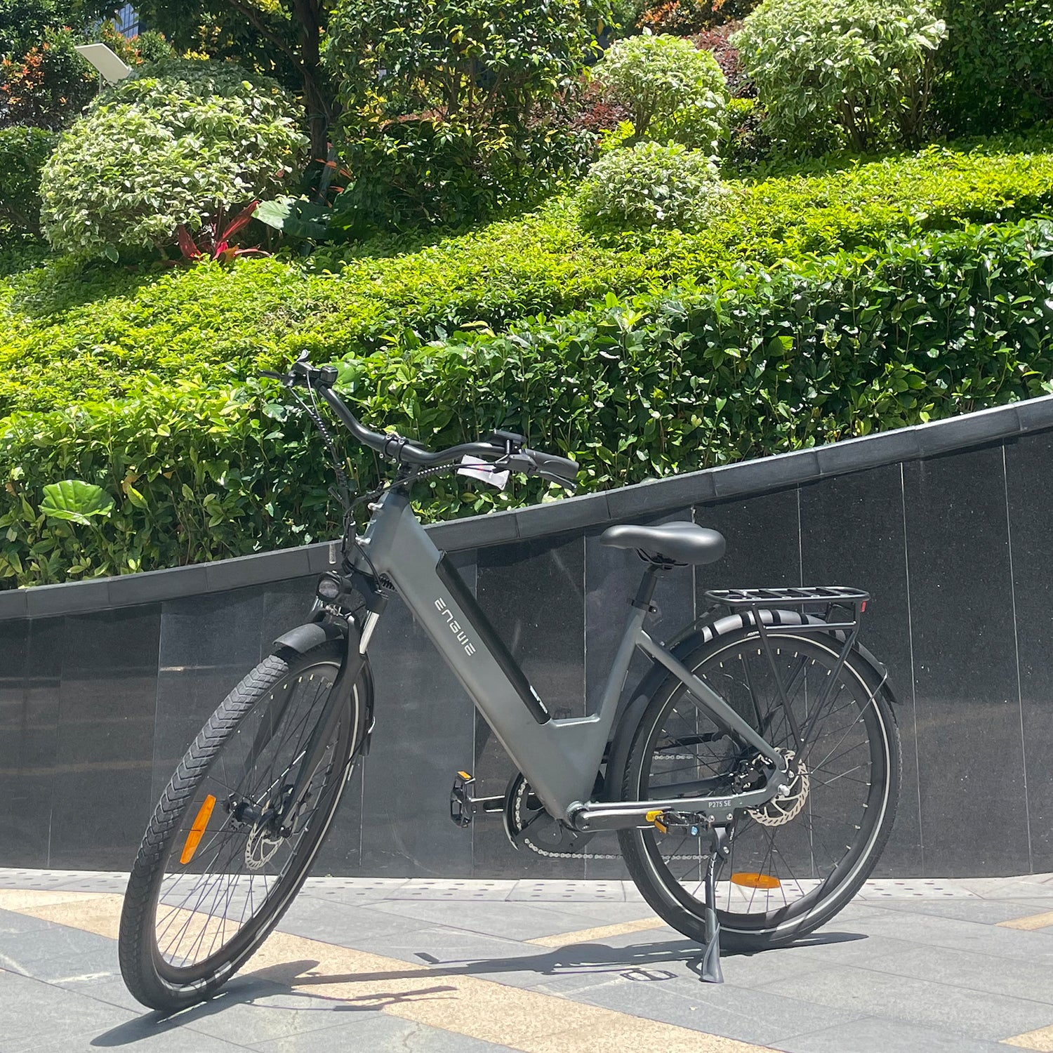 Una bicicleta eléctrica ENGWE gris estacionada en una acera. Frondosos arbustos y árboles verdes en el fondo.
