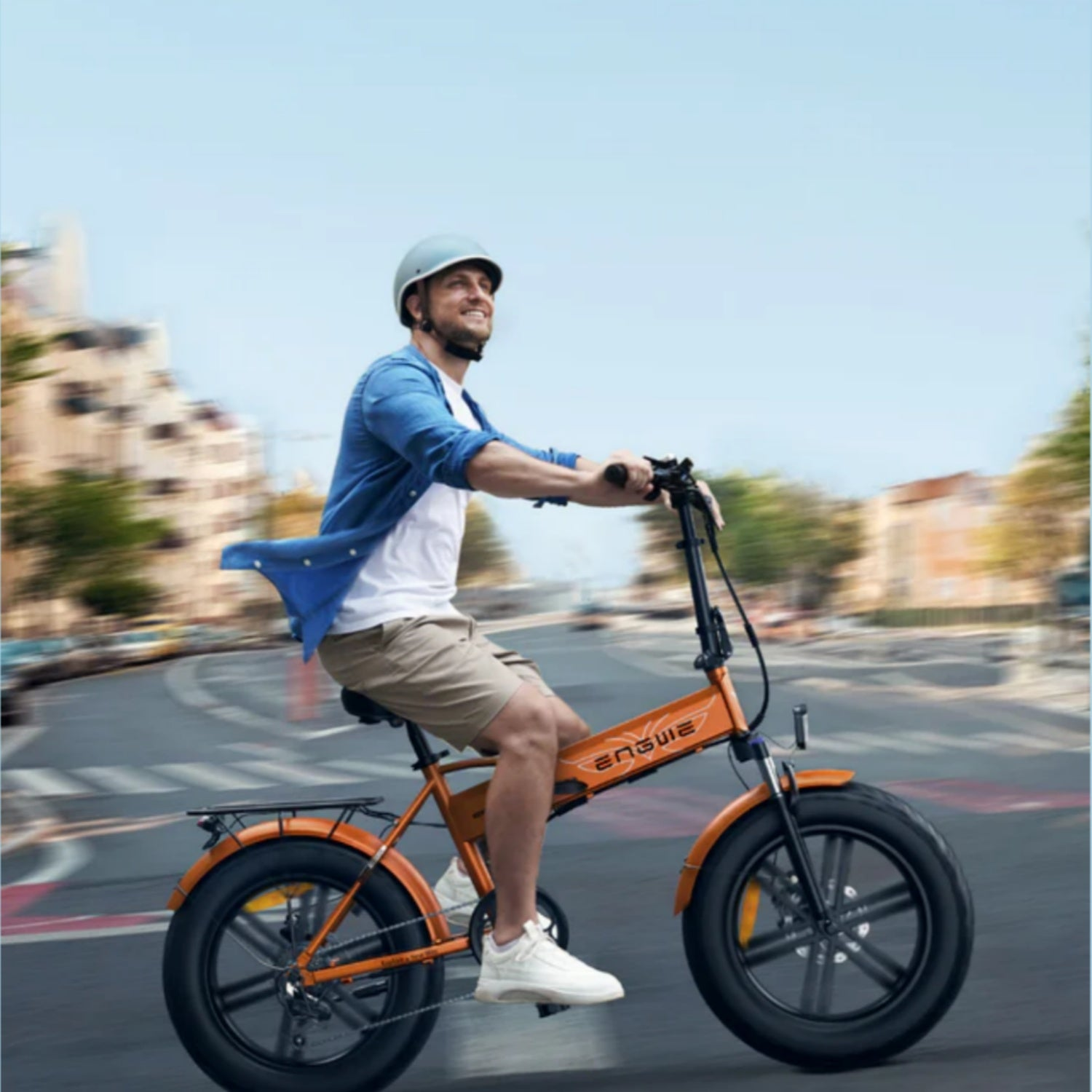 Un hombre monta en una bicicleta eléctrica naranja en una carretera, con casco y mirando hacia arriba. Edificios y árboles al fondo.