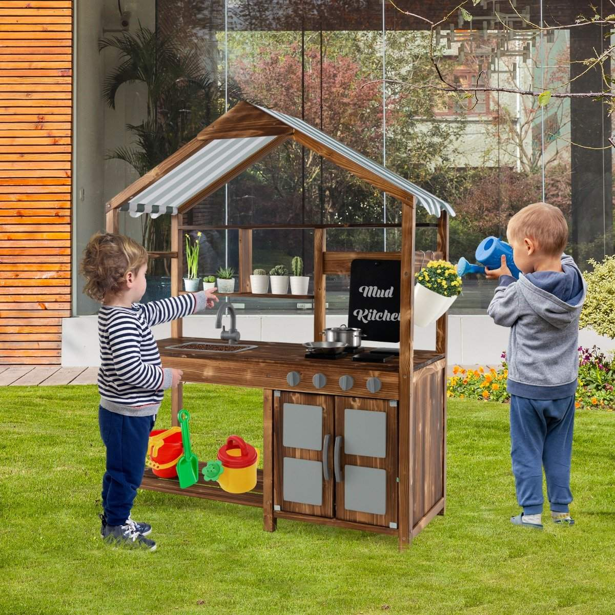 Dos niños juegan con una cocina de barro en un jardín. Uno señala, el otro riega flores con una regadera azul.