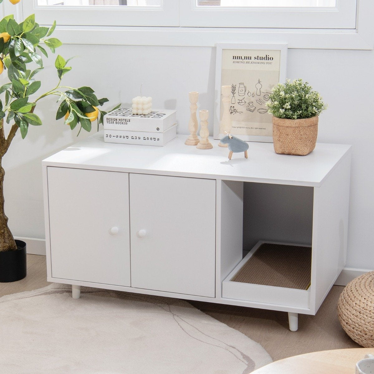 Muebles blancos para gatos con puertas cerradas, estante abierto. Decoraciones incluyen plantas, libros, velas y un cuadro enmarcado. Un limonero a la izquierda.