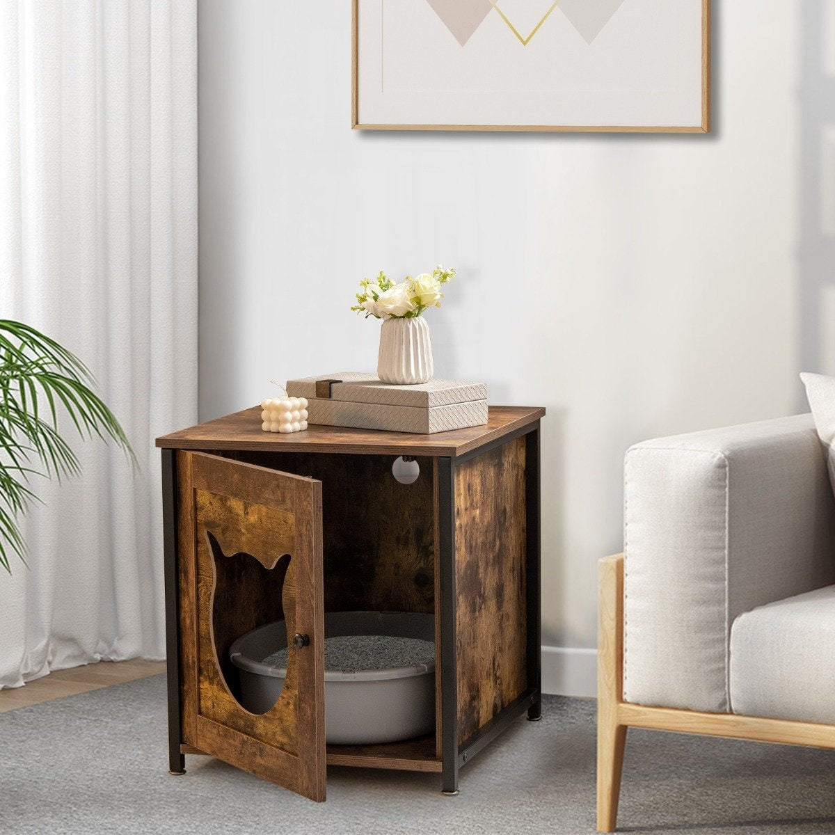 Un arenero para gatos dentro de un mueble de madera con puerta en forma de gato. Flores y decoración encima. Un sofá y una obra de arte al fondo.