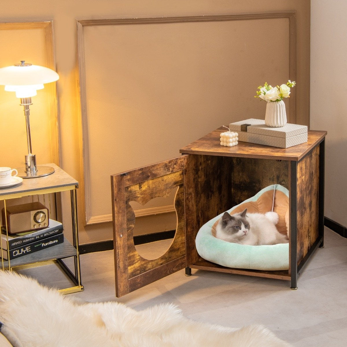 Un gato descansa en una cama dentro de una casa para gatos de madera. A la izquierda se ven una lámpara y una mesita auxiliar. Flores en la parte superior.