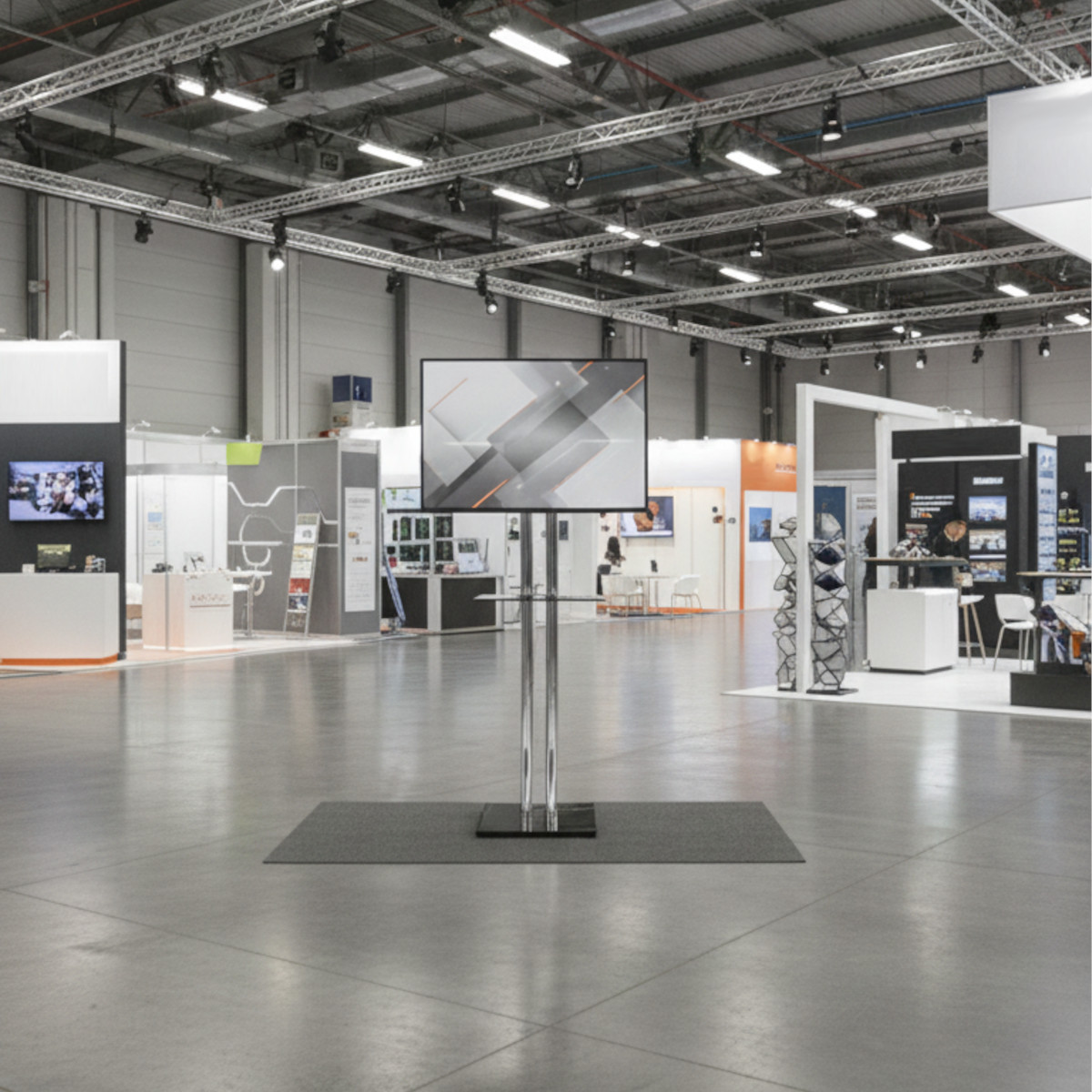 Eine leere Ausstellungshalle. Ein großer Bildschirm zeigt ein geometrisches Design. Stände und Personen sind im Hintergrund. Der Boden ist grau.