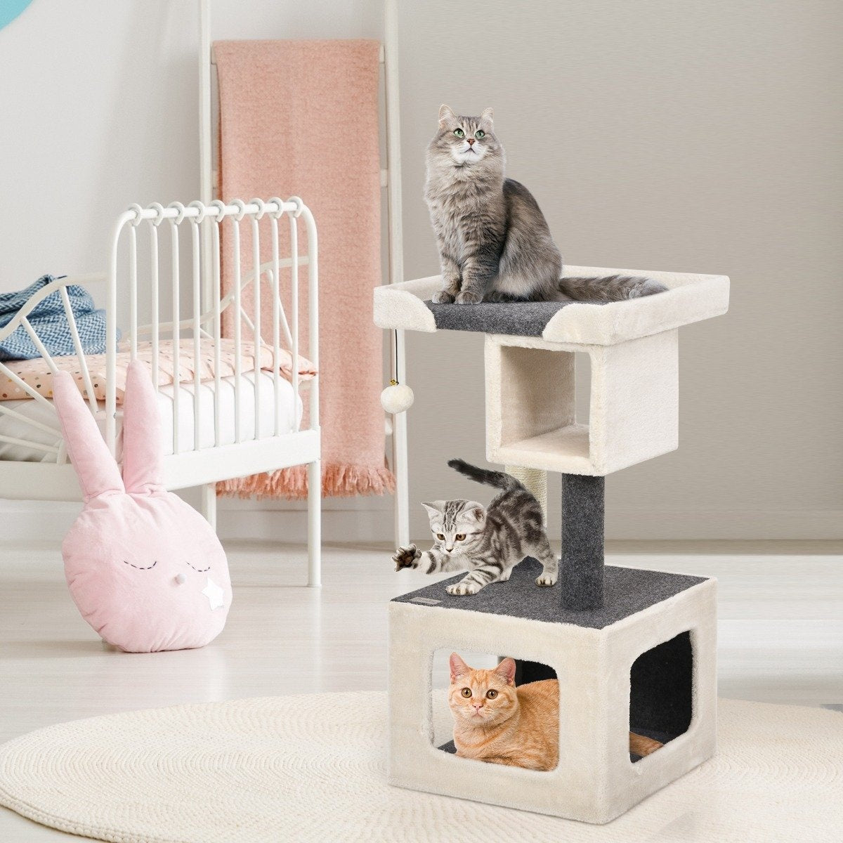 Tres gatos en un árbol para gatos. Uno arriba, otro juega y el tercero descansa dentro. Almohada de conejo rosa y cuna blanca al fondo.