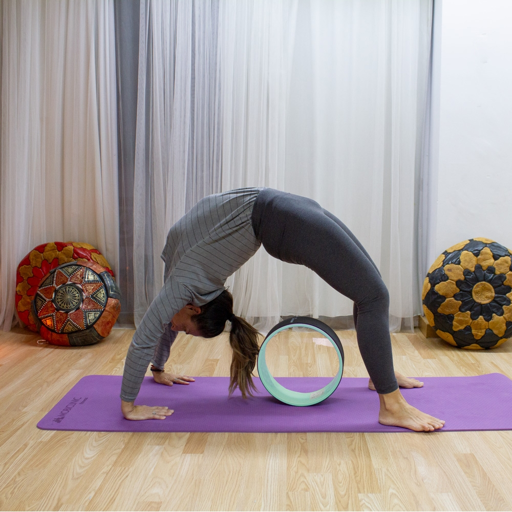 Mujer en pose de puente con rueda de yoga sobre esterilla morada. Cojines decorativos al fondo. Suelo de madera y cortinas blancas.