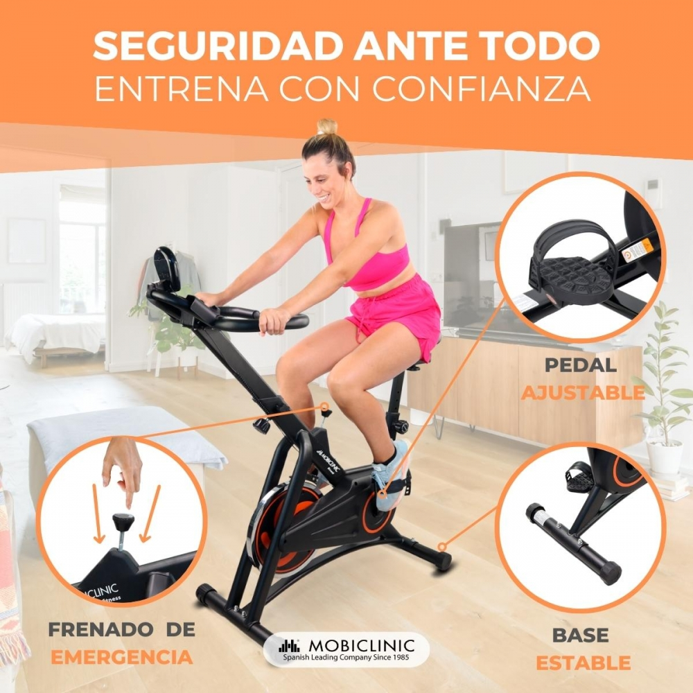 Mujer en ropa deportiva rosa en bicicleta estática. Detalles muestran pedal ajustable, freno de emergencia y base estable. Fondo es una habitación.