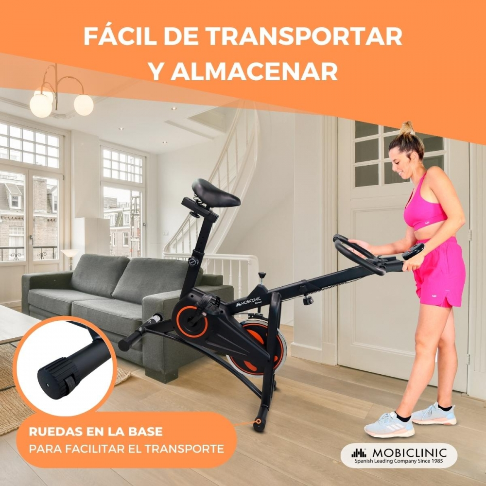 Una mujer en ropa deportiva rosa usa una bicicleta estática. La bicicleta es negra con detalles naranjas. Ruedas en la base.