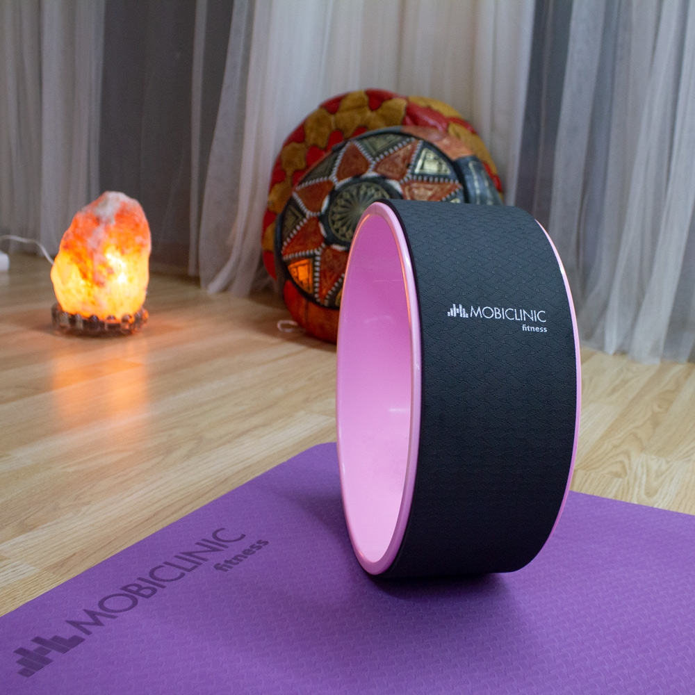 Una rueda de yoga con exterior negro e interior rosa está sobre una esterilla morada. Una lámpara de sal y una almohada decorativa son visibles al fondo.