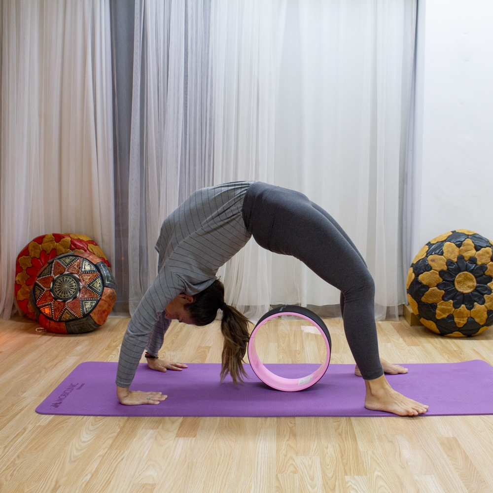 Una mujer en pose de yoga, con la espalda arqueada. Usa una rueda en una esterilla morada. Cojines decorativos a ambos lados.