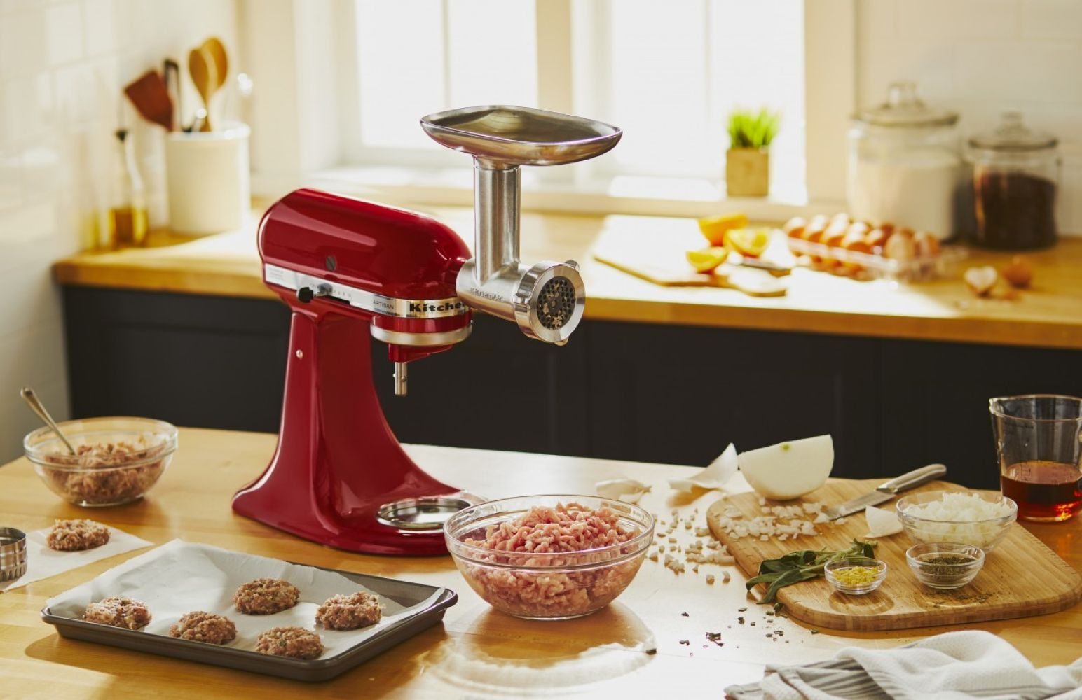 Ein roter KitchenAid-Fleischwolf mit Fleisch, Zutaten und fertigen Patties auf einer Küchentheke. Im Hintergrund ein Holzschneidebrett und ein Fenster.