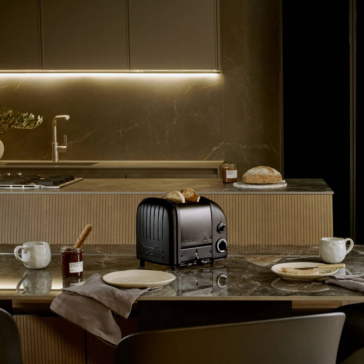 Ein schwarzer Toaster mit Brotscheiben steht auf einem Tisch mit Teller, Marmelade und Becher. Ein Brotlaib liegt im Hintergrund.