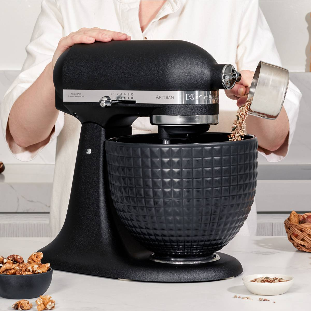 Eine Person gießt Getreide in eine schwarze KitchenAid-Mixerschüssel. Walnüsse und andere Zutaten liegen auf der Theke.