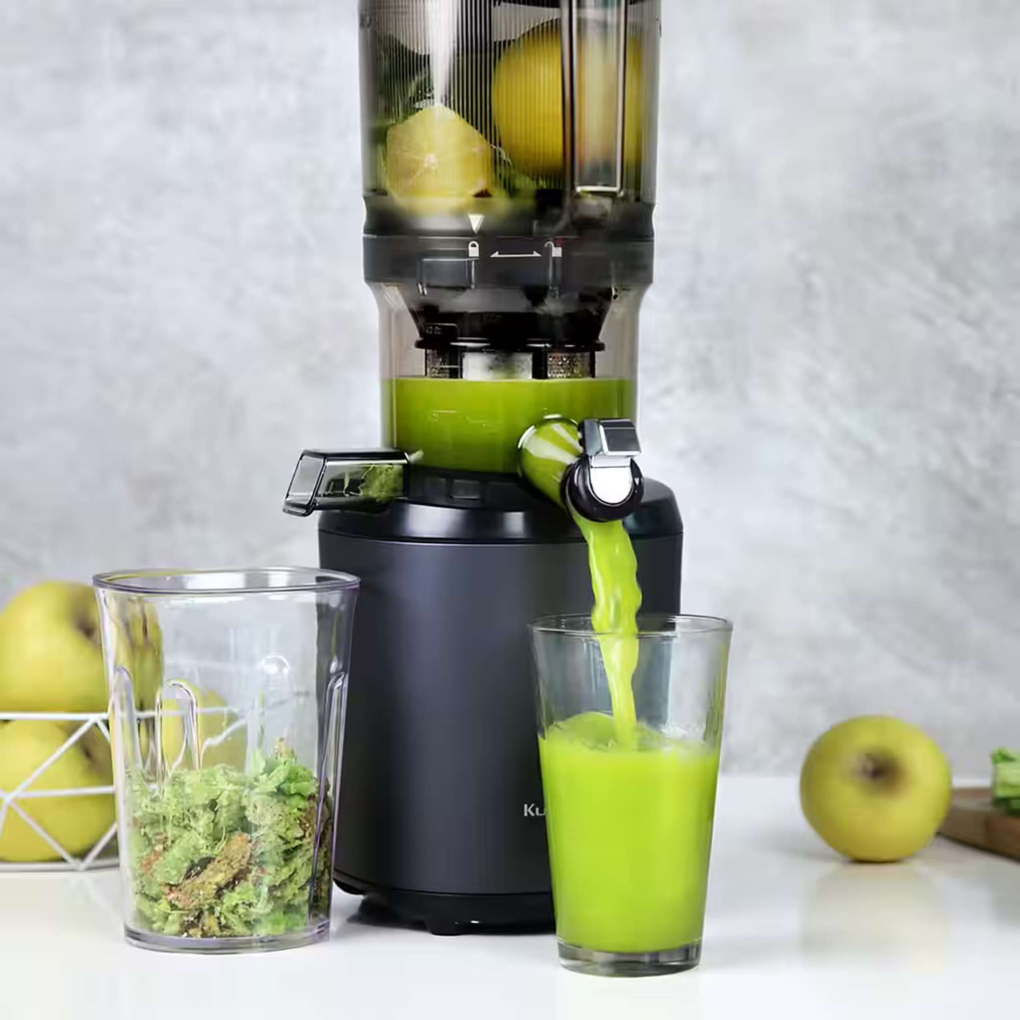 Ein Entsafter extrahiert grünen Saft in ein Glas. Zitronen sind im Entsafter. Äpfel und Fruchtfleisch sind sichtbar.