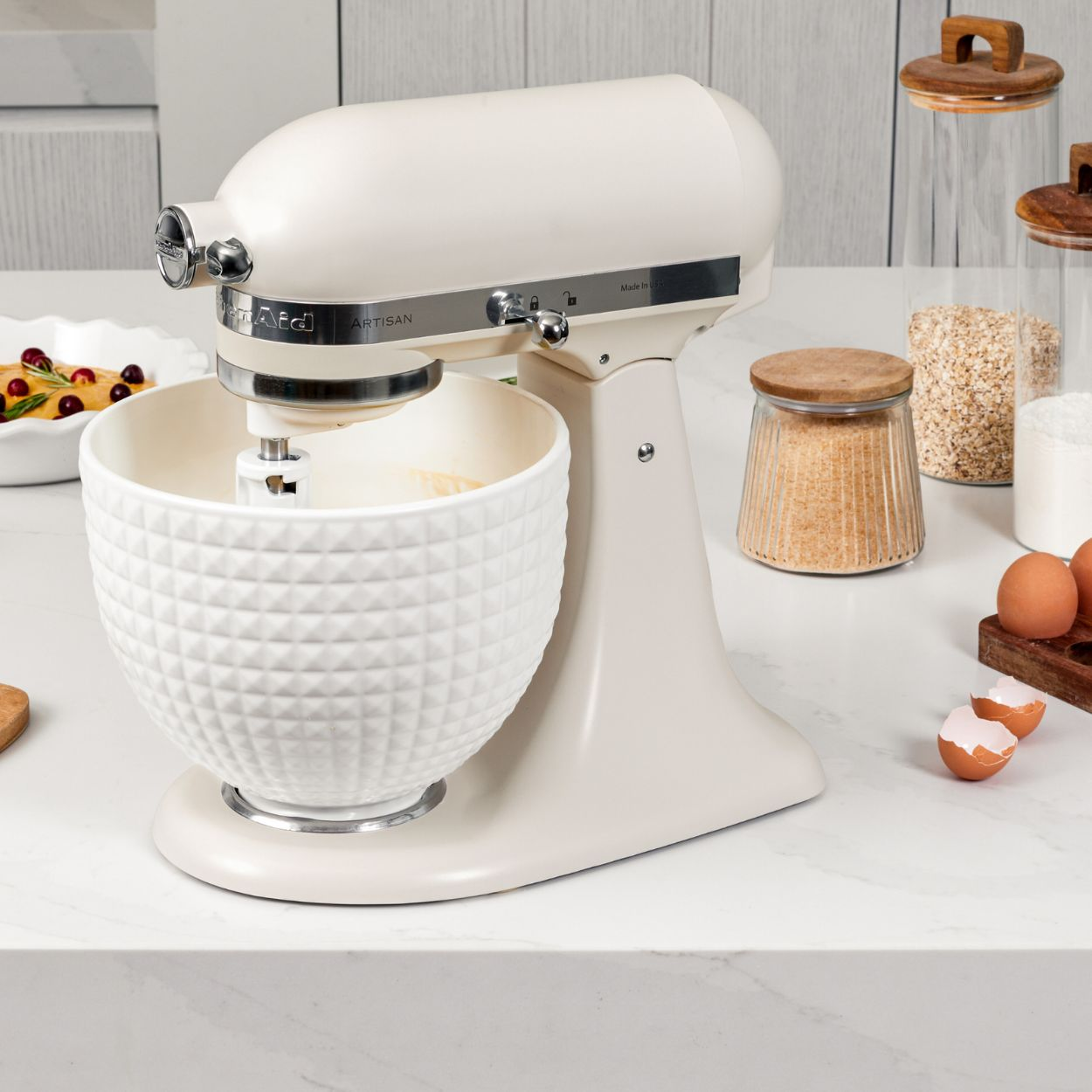 Cremefarbener KitchenAid-Mixer mit weißer, gemusterter Schüssel. Gläser mit Zutaten, Eier und eine Schale mit Beeren stehen auf einer Marmorplatte.