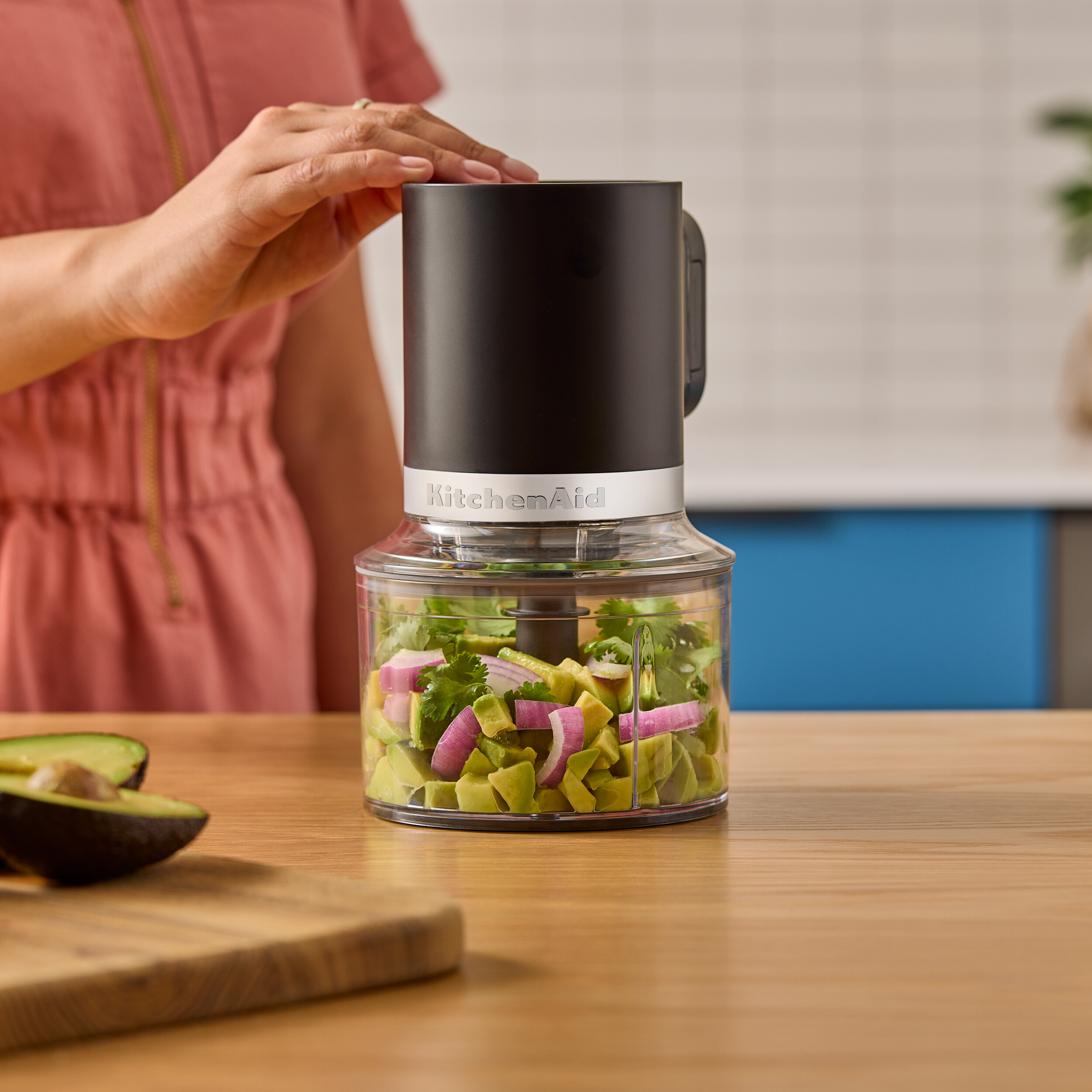 Ein Foodprozessor mit Avocado, roter Zwiebel und Koriander. Die Hand einer Frau liegt auf dem Deckel. Avocadoscheiben daneben.